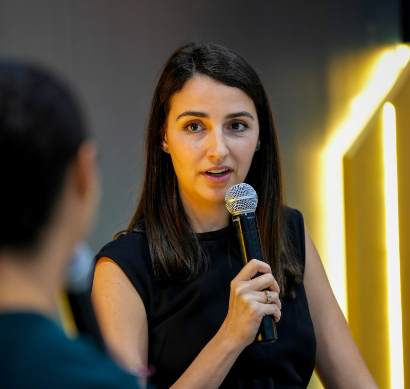 Two women engaged in conversation while seated on stage, sharing insights in a professional setting.